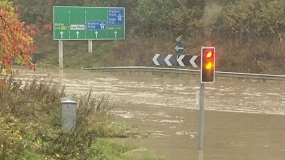 Brown water flowing around a roundabout off the M62 motorway, with traffic lights at a junction changing from red to green.  