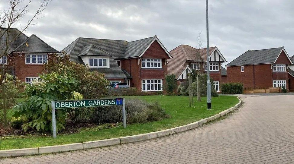 A wide view of Oberton Gardens, with a sign on a lawned area with bushes, and a number of detached houses visible.