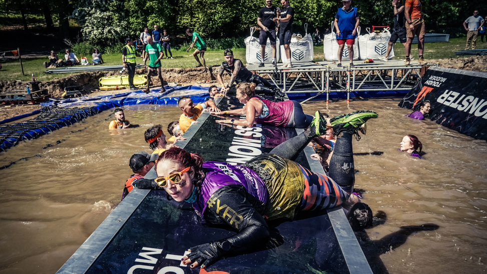 Debbie in an event top and leggings, climbing out of a mud bath onto a platform, grabbing on with her hands. She's surrounded by other participants, some of whom are in a bath of mud up to their necks