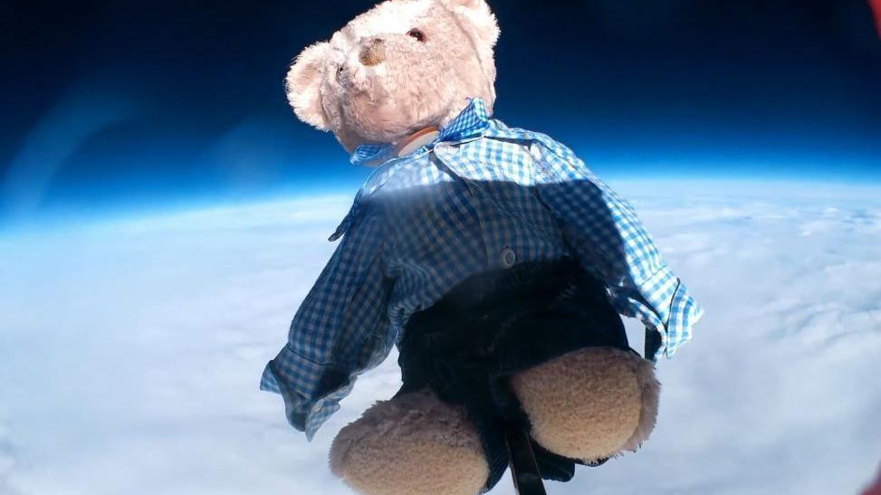 Small, brown bear, wearing school uniform, navy blue shorts and a blue and white checked shirt flying attached to a weather balloon above clouds