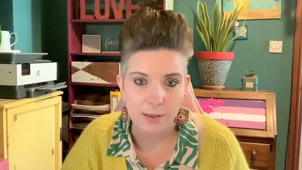 A woman in a yellow jumper sat in front of a green wall.