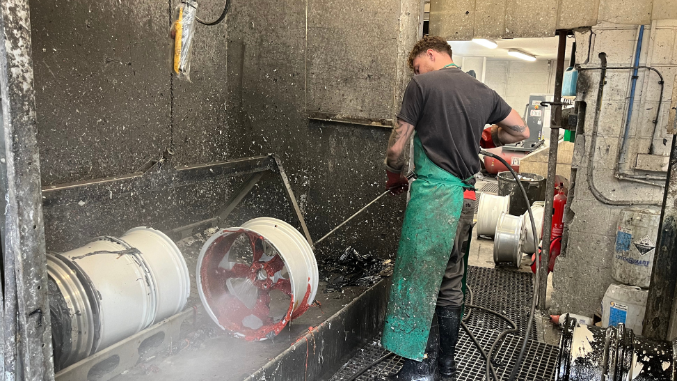 Keiran Thomas strips old alloy wheels. He is photographed from the side and he is wearing a green apron and black T-shirt and is working in a garage. 