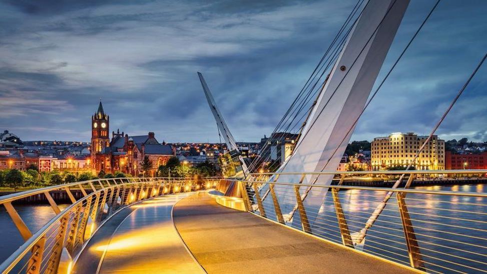 Londonderry's peace bridge spanning the foyle, with a view of the city's Guildhall to the left and the city hotel to the right. It is dusk, and the sky is overcast, the bridge and buildings are lit up