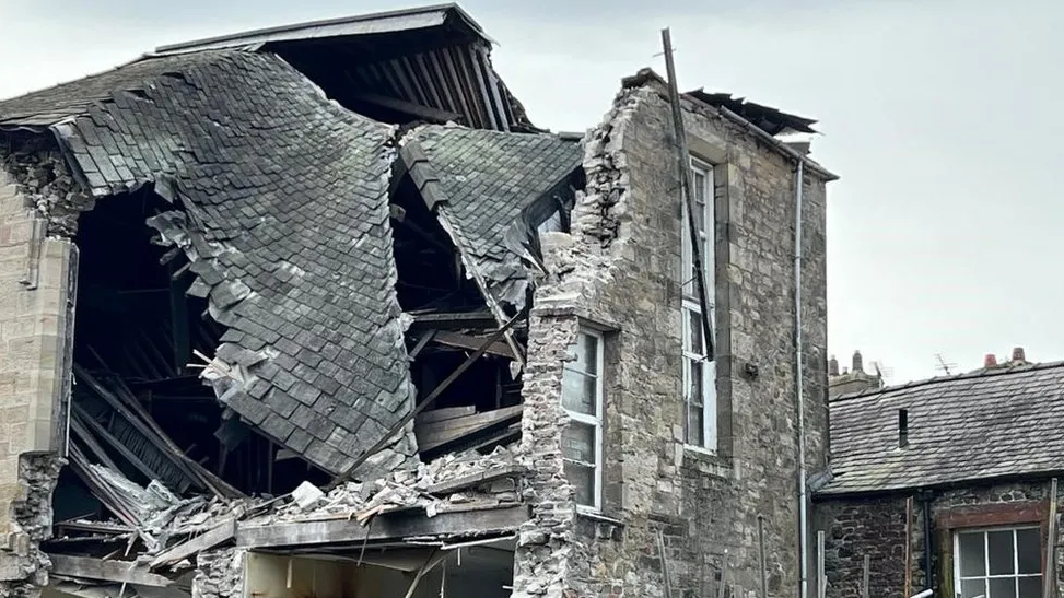 A grey-stoned three-storey building that has collapsed. A wall is missing with brickwork and metal poles hanging precariously.