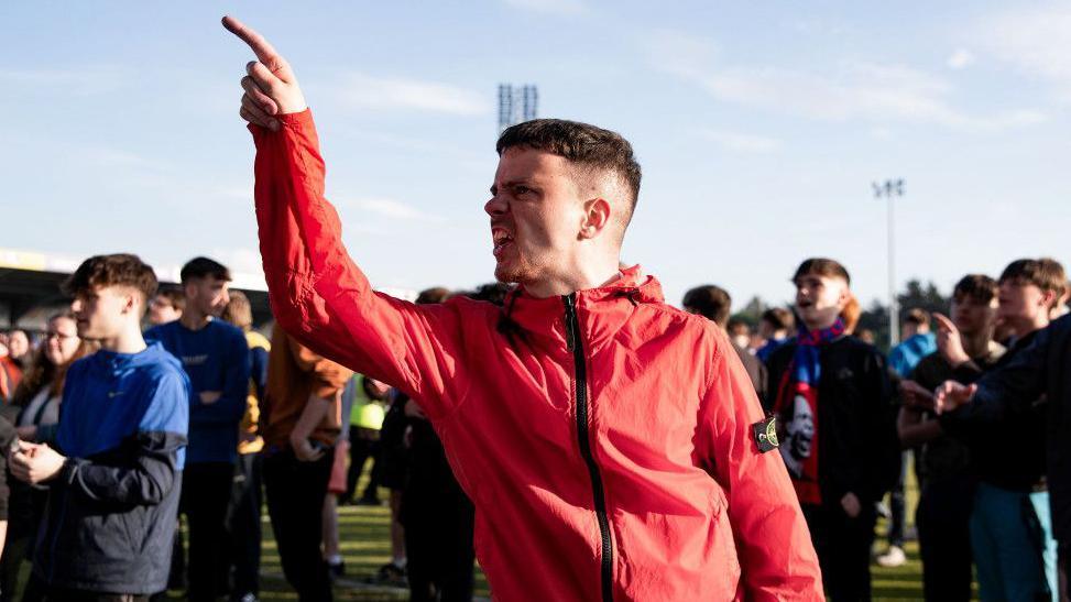 An Inverness fan wearing a red jacket and standing in a crowd of fellow ITC supporters points angrily after the club was relegated.