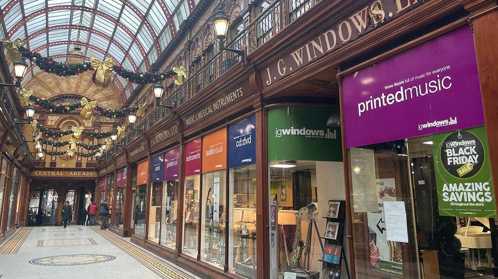 The multi-coloured shop front of J.G Windows. Musical instruments can be seen through the windows. Christmas decorations are hung in the arcade hallway outside.