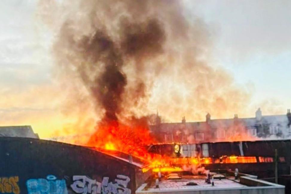 Flames in a building with smoke rising into the sky, with buildings in the background.