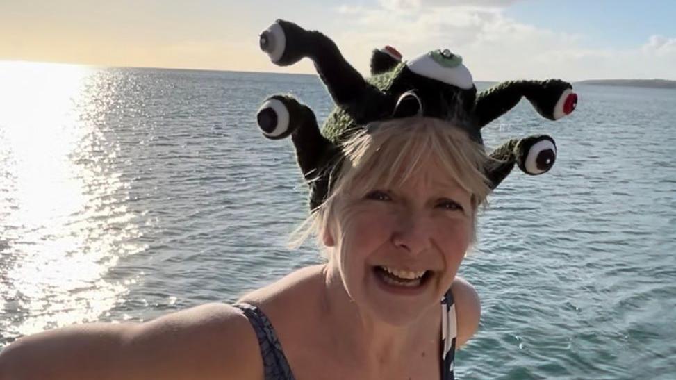 Ruth standing smiling by the sea wearing a swimming costume and a green and white knitted hat topped with 3d eyeballs. 