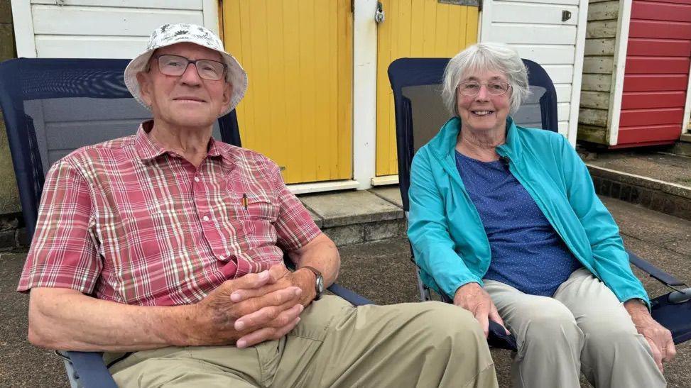 Colin and Doreen Hunt sat in seaside fold out chairs outside of a white beach hut with yellow doors. Colin, on the left, is wearing a red and white flannel shirt and grey trousers. Doreen is wearing a blue shirt, a blue zip up jacket and grey trousers. 