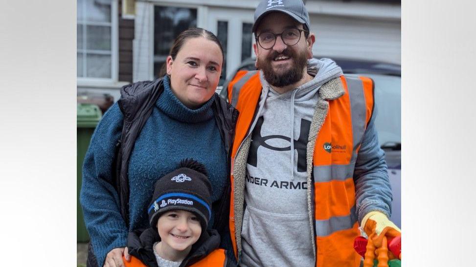 Harlee is standing with his mum and dad. He is wearing a black and blue beanie hat with a black and grey coat and an orange hi-vis jacket. His mum has brown hair and is wearing a blue jumper with a black gilet. His dad has brown facial hair and is wearing a grey cap, grey hoodie and orange hi-vis jacket. He is wearing gloves and holding the top of some litter collection tools