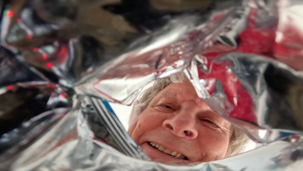 A photo taken from inside the silver wrapper of an open crisp packet. Frank Munford's face is towering above as he looks in through the opening. He has grey hair and a cheery smile.