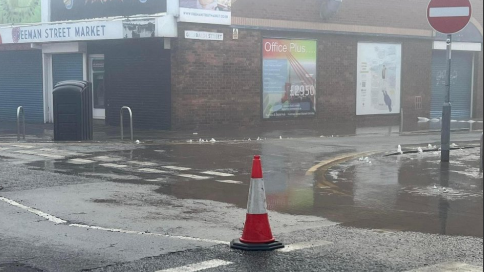 Flooding on street in Grimsby - a lone traffic cone stands at the edge of a large pool of water. Freeman Street Market hall is next to the pool of water. 