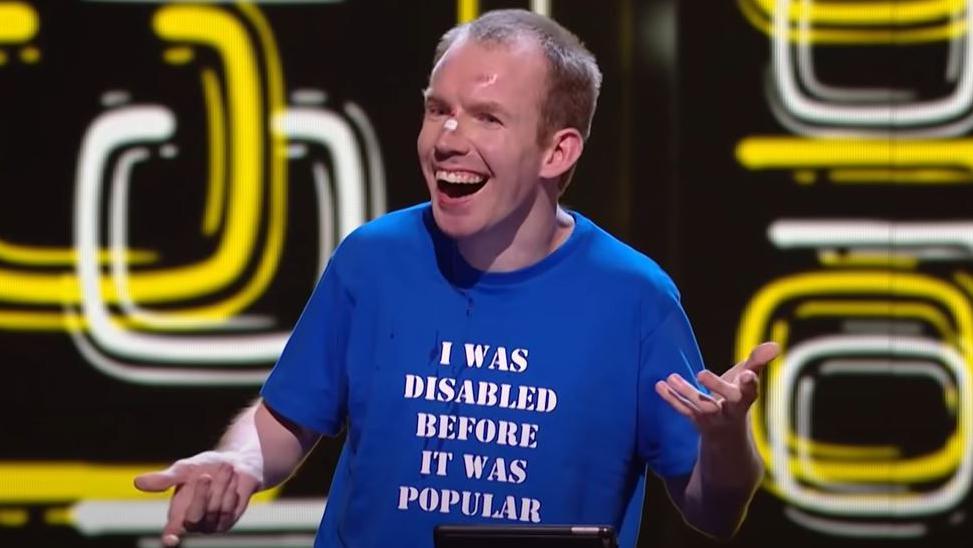 A man on stage wearing a blue T-shirt with the slogan "I was disabled before it was popular"