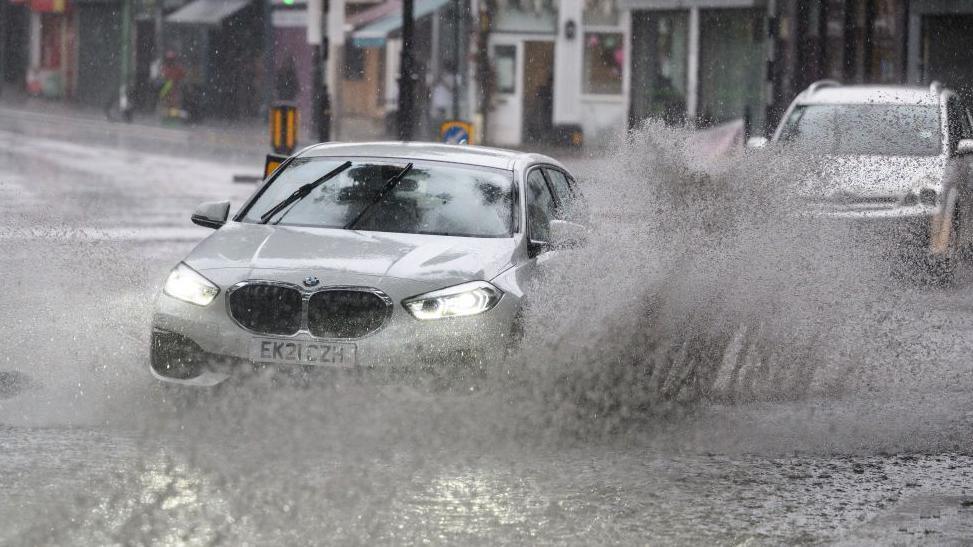 A silver car drives through water on the road in suburban London, creating a large splash.