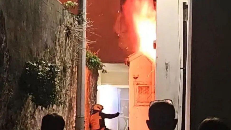 A firefighter inspects the outside of a building which is on fire with flames billowing from the roof. Three people standing at the edge of a cordon can be seen watching on. A large stone wall is to the left of the image.