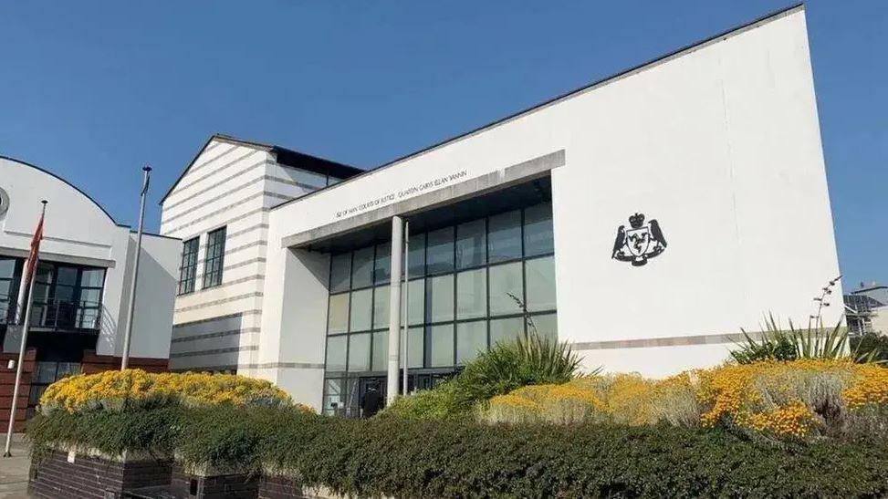 The Isle of Man Courts of Justice - a large, white glass-fronted building with the government crest on the right-hand side. There are planters leading to the entrance, featuring green and yellow shrubbery.