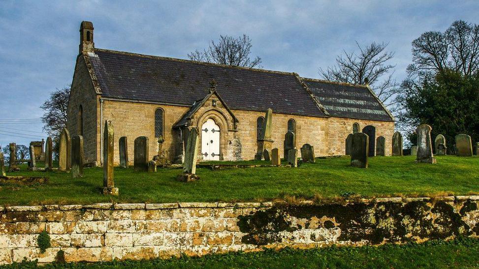 A small church with a white door. It sits in a graveyard, in a raised area with a wall in the foreground of the image.