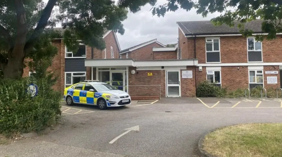 A police car parked outside a red-brick, double-storey housing complex. There is a car park to the front of the building and a green patch of grass.
