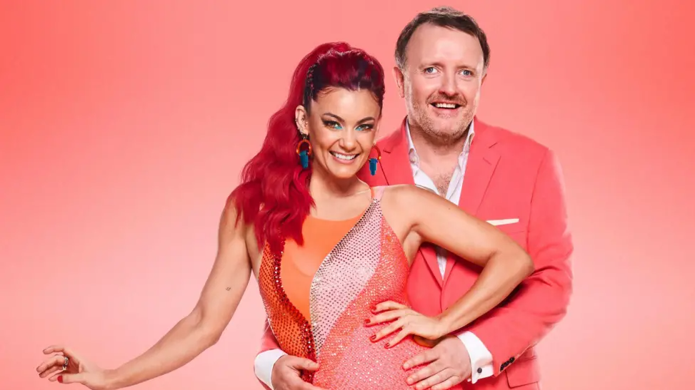 Comedian Chris McCausland, wearing an open-necked white shirt and a pink jacket, holds the waist of dancer Diane Buswell, who has long dyed bright red hair and who is wearing a brightly-coloured sparkly leotard, in a promotional shot.