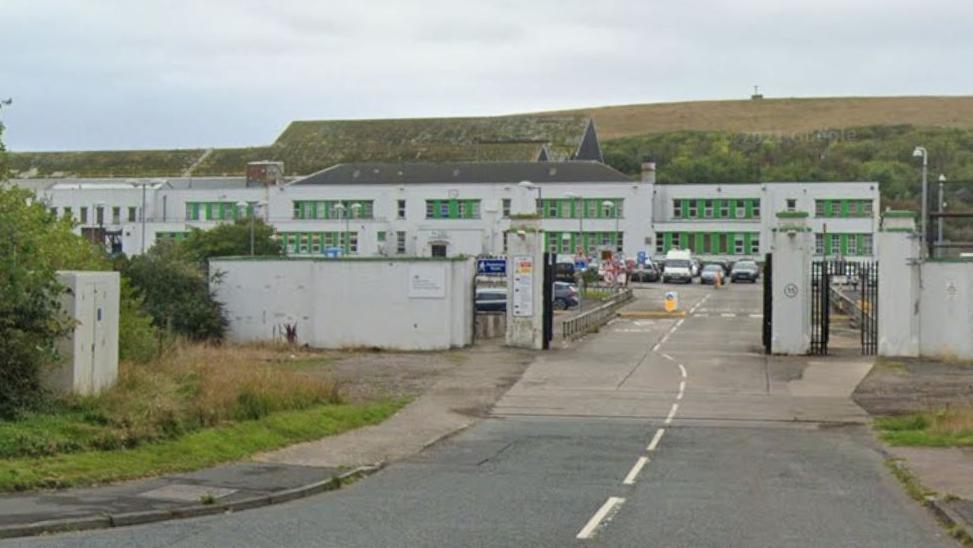 A street view of the Chapel Bank Engineering building in Workington. It's a large white building with green windows. There's a road and car park in front of the building and a large hill behind it.