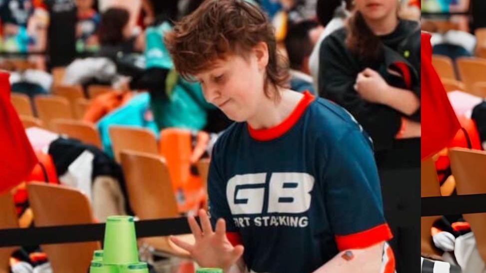 Laura Beacom has red hair and is stacking green cups on a table. She is looking down at the table and has her hands poised above the cups. She is wearing a dark blue sports top which reads "GB sport stacking" in large white letters. There are chairs in the background and people standing around.