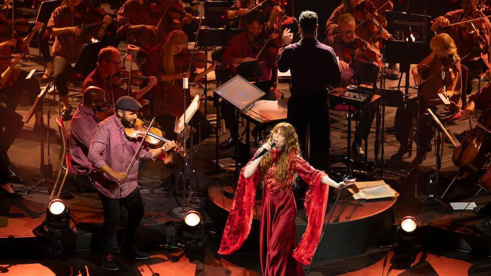 Florence stands in front of an orchestra wearing a long silky red dress with huge wing-like sleeves, as she sings into a microphone. She has long auburn wavy hair and her left arm reaches out and holds the mic stand. Jules is seen from behind, conducting, and in the orchestra we can see mainly strings players who are dimly lit.