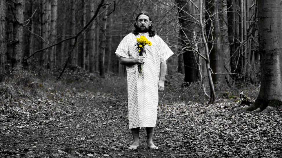 Lou Chawner is stood in the middle of the image in a forest. He is wearing a long medical gown and is holding a bunch of yellow flowers in his right hand. He has shoulder length hair and is looking towards the camera.