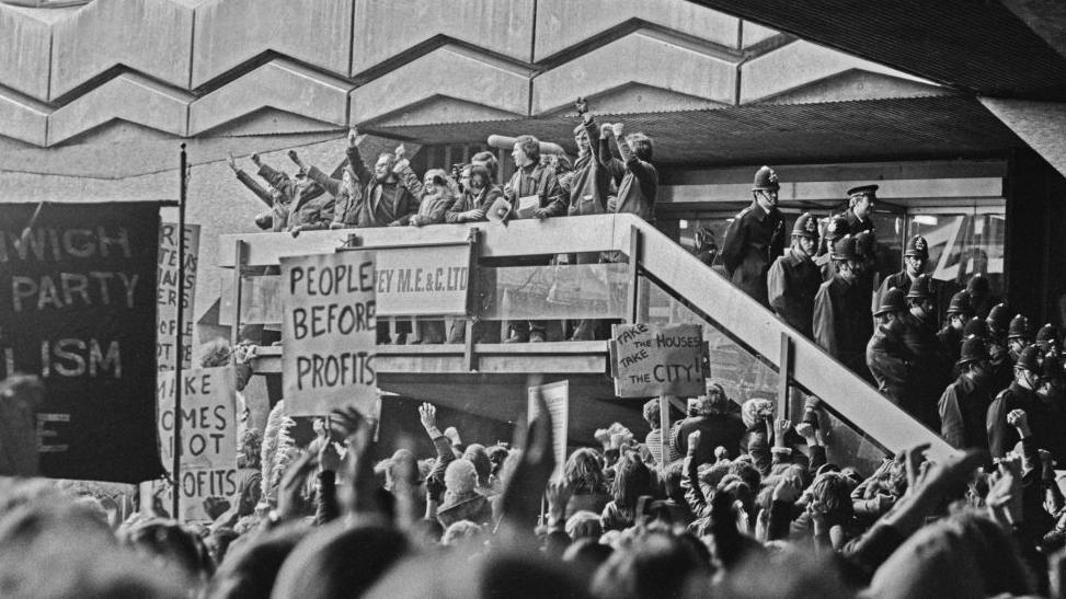 A black and white picture of a protest. There are around 10 people standing on a balcony, some with their fists in the air. On the floor below, there are around 100 people looking up. Some are holding signs, with slogans reading: 'People before profits' and "Take the house, take the city'. There are also around 15 police officers standing on a staircase. 