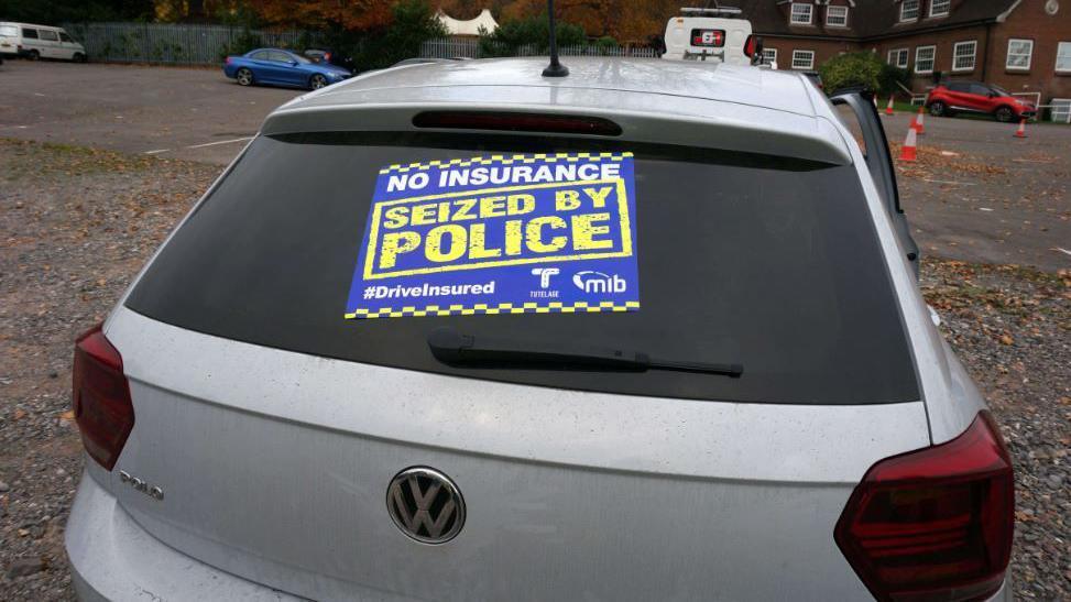 A rear shot of a white Volkswagen with a large sticker across the rear windscreen which reads 'no insurance, seized by police" 