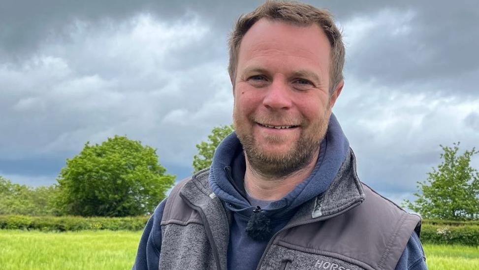 Olly, wearing a blue hoodie and grey gilet, is standing in the middle of a field on a cloudy day.