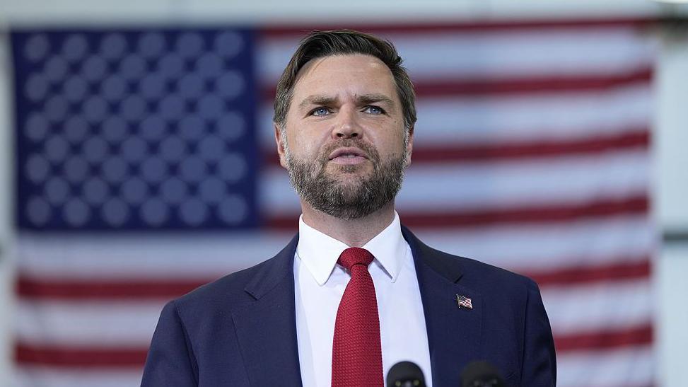 A man in a suit. He is wearing a red tie and has the US flag pinned on his lapel. Behind him is the US flag.