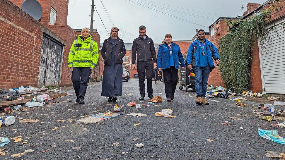 A police officer in a hi-vis jacket walking besides Irim Ali, who is wearing a dark jacket and long dress, an environmental protection manager and two community safety support officers who are wearing bright blue tops and jackets. The street is covered in rubbish.