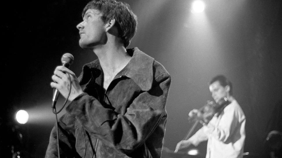 A thin man in a jacket which looks a bit too big for him hold a microphone and looks into the middle distance