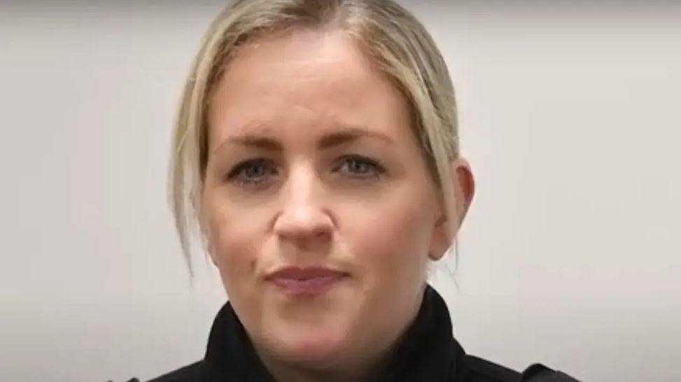 Female police officer with blond hair and wearing a dark jacket stares at the camera