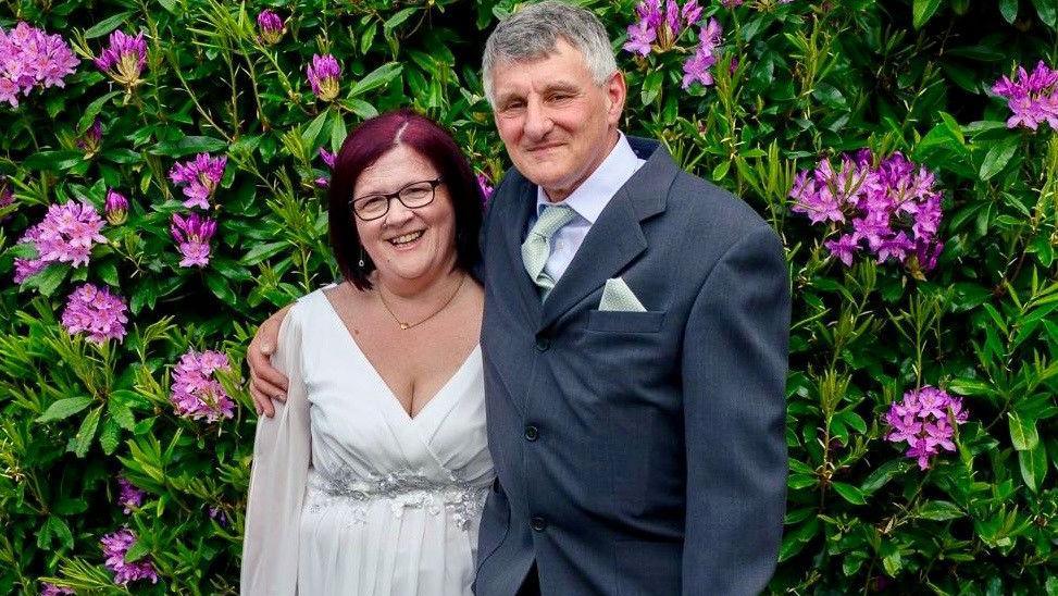 Couple on their wedding day with a black Labrador in front of them. The woman is wearing a white dress and the man is wearing a suit.