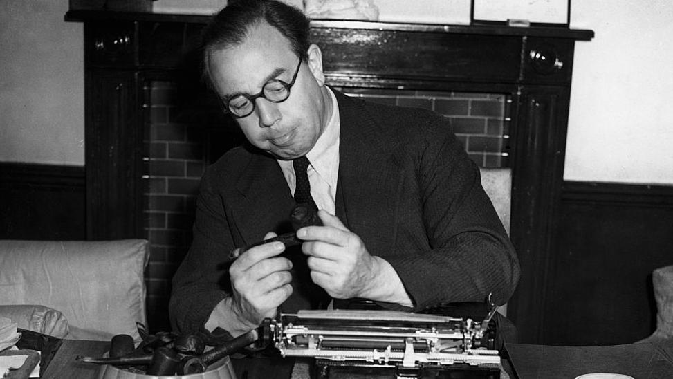 A black and white image of the author J B Priestley - a man in a dark suit and tie and white shirt, wearing dark-framed glasses, with combed back dark hair - who is inspecting a pipe he is holding with both hands. He is sat at a desk with a typewriter on it