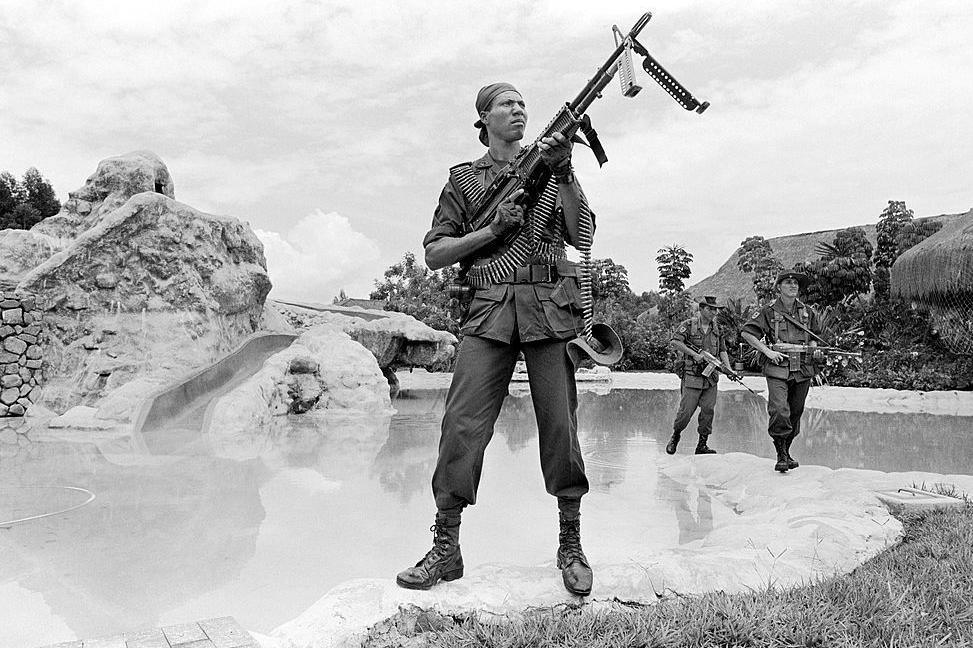 In a black and white photograph soldiers carry heavy weapons are they search a compound.