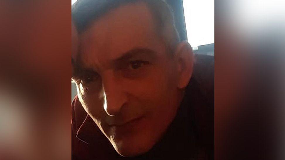 Paul Foster with brown hair and wearing a red jacket. There is a window behind him letting in daylight.