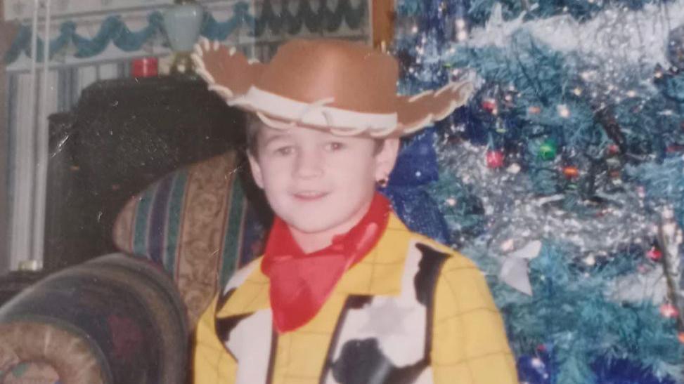 A photograph of Kaine Fletcher as a child, wearing a Woody costume, from Toy's Story', standing in front of a Christmas tree