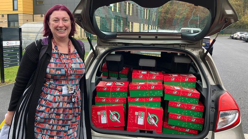 Careers adviser Jo Kerr stands next to a car with red and green shoeboxes loaded up in the boot. She is wearing a dress and black cardigan and a lanyard.