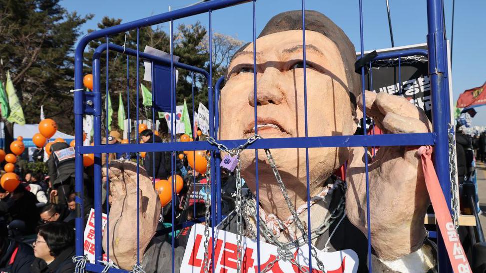 A sculpture of South Korean president Yoon Suk Yeol is draped in chains inside a blue cage and surrounded by protesters in the street