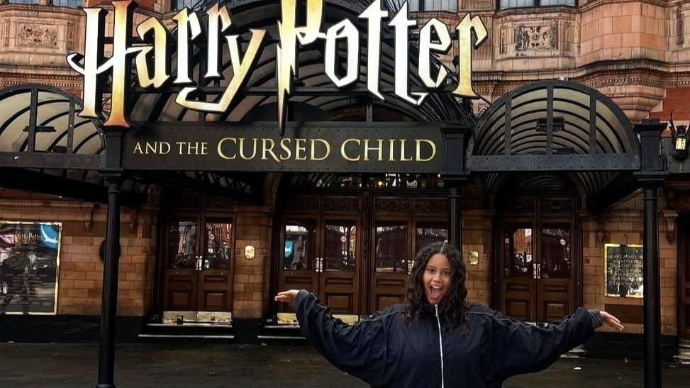 Mariam Pope wearing a black jacket while standing in front of the Palace Theatre with the Harry Potter and the Cursed Child logo behind her. She is smiling and holding her hands out.