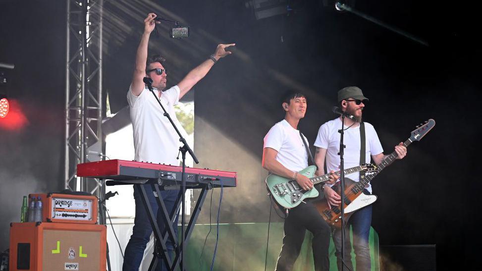 Scouting for Girls on stage - frontman Roy Stride has his arms raised above his head, with the other band members playing  guitars