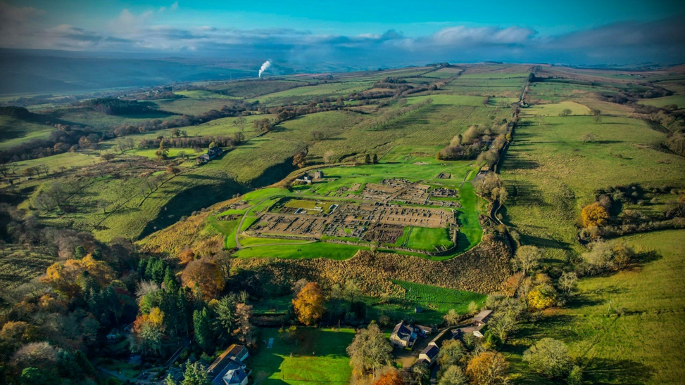 An aerial shot of the Vindolanda Fort which stretches over a large site. Beyond it the fields of the Tyne Valley stretch into the distance 