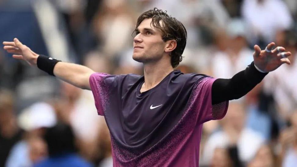 A white man with long brown hair with his arms raised by his side. He is wearing a pink and purple Nike sports shirt and is looking up.