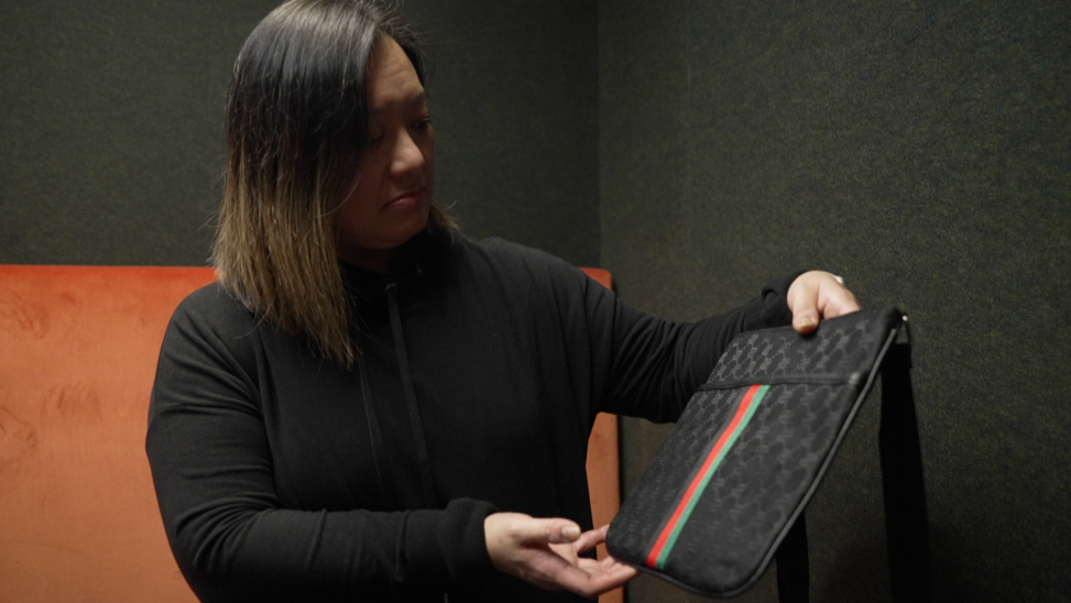 A woman holding a black bag, it has an interlocking design and black,red and green stripes down the centre. The woman is wearing a black hoodie and has medium length hair.