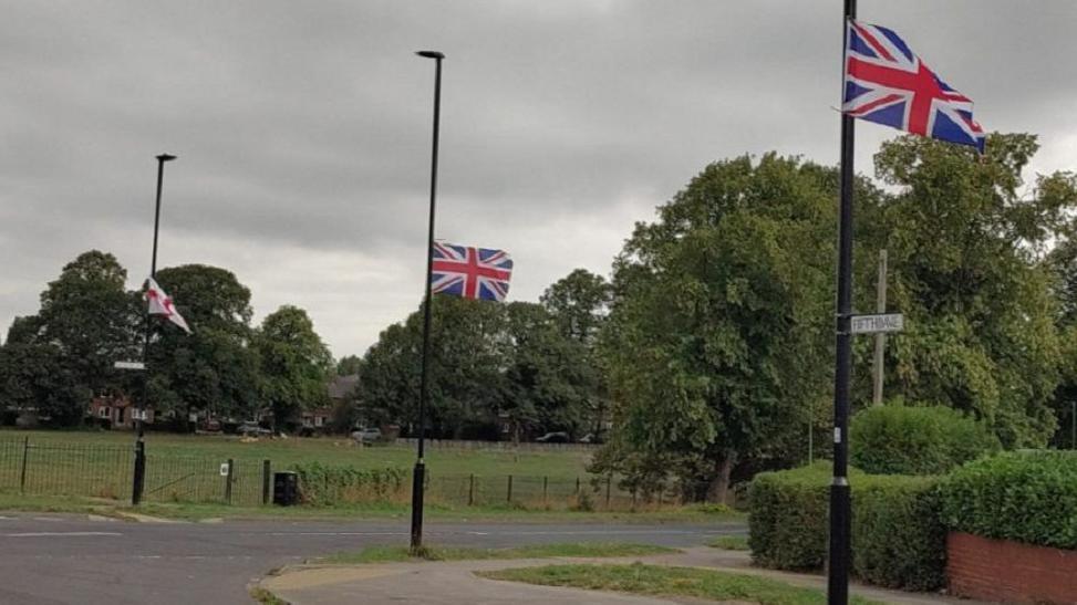 Union jack flags placed on lampposts in a street that has greenery and bushes and trees in the background