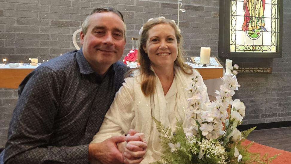 Emily and Jamie Cross in the chapel at the Royal Preston Hospital. They are smiling and holding hands sitting in front of a stained glass window. She is wearing white pyjamas and is holding white flowers with green foliage. he is wearing a dark grey smart shirt