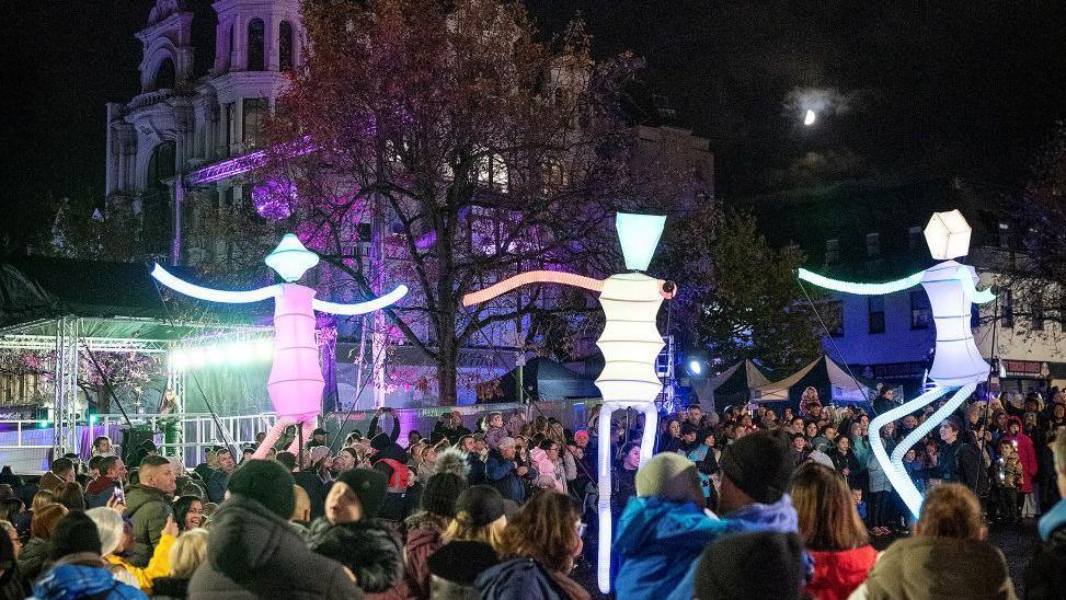 Shows large light up puppets dancing with lots of people in the background