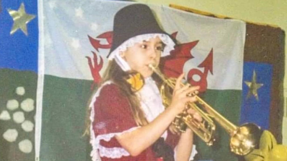 An old image of Lauren playing the trumpet in a St David's day costume on stage. She is standing in front of a Welsh flag.